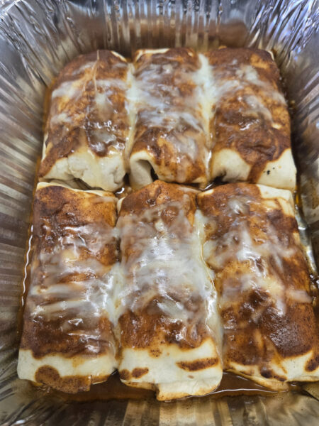 A tray of baked Breakfast Enchiladas, featuring six enchiladas covered in melted cheese and Tex Mex sauce, resting in a savory sauce. The enchiladas are golden brown and arranged neatly in a disposable aluminum tray.