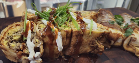 Close-up of Breakfast Enchiladas topped with green onions, sour cream, and Tex Mex sauce, served on a wooden board. The enchiladas are filled with breakfast sausage, scrambled eggs, and Monterey Jack cheese.