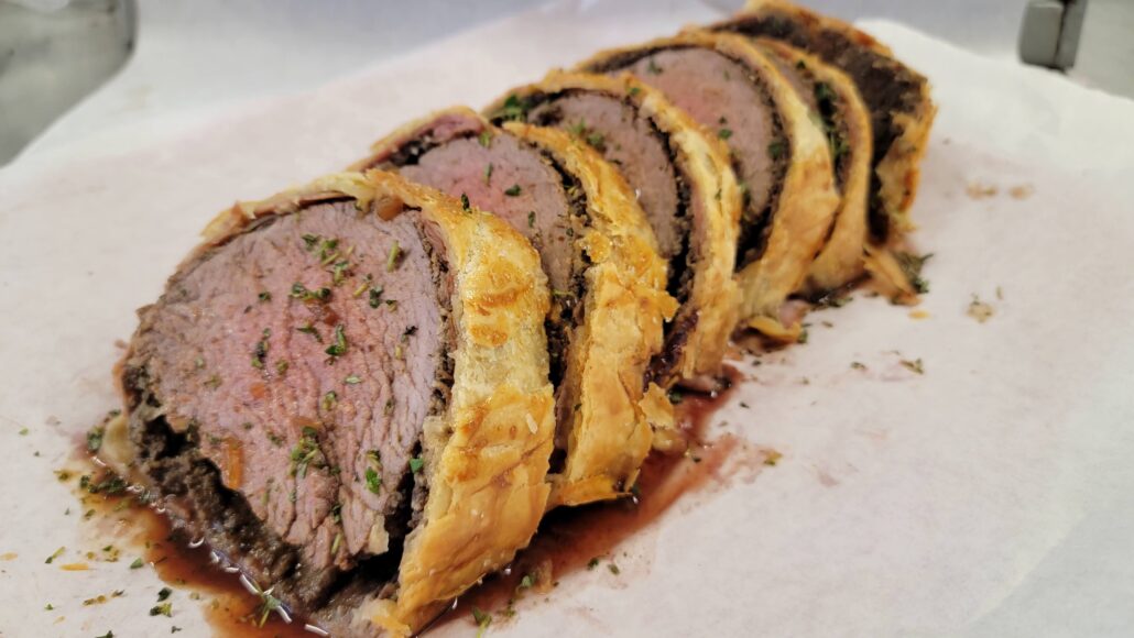 Sliced Beef Wellington on a white parchment paper, showcasing tender beef fillet wrapped in flaky golden pastry. The dish is garnished with herbs and accompanied by a rich sauce.