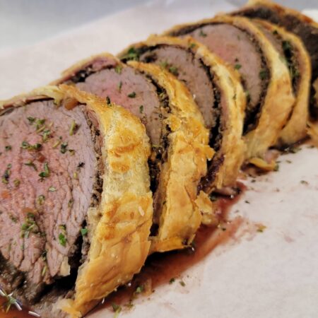 Sliced Beef Wellington on a white parchment paper, showcasing tender beef fillet wrapped in flaky golden pastry. The dish is garnished with herbs and accompanied by a rich sauce.
